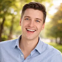 Ein junger Mann mit kurzen braunen Haaren, der ein hellblaues Button-up-Hemd trägt, lächelt breit, während er im Freien in einem sonnenbeschienenen Park mit Bäumen im Hintergrund steht.