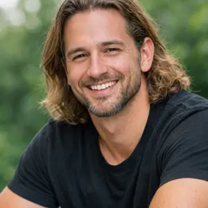 Ein Mann mit schulterlangen braunen Haaren und einem Bart lächelt in die Kamera, er trägt ein schwarzes T-Shirt, vielleicht kurz nach dem Abschluss eines GmbH Verkauf Notar-Termins, mit einem unscharfen grünen Hintergrund im Freien.