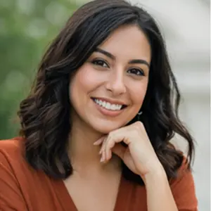 Lächelnde Frau mit schulterlangen dunklen Haaren, die ein orangefarbenes Top trägt, stützt ihr Kinn auf ihre Hand. Der unscharfe grüne Hintergrund deutet eine Umgebung im Freien an, ideal für kreative Gedanken oder die Planung einer GmbH gründen in Berlin.