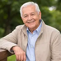 Ein älterer Mann mit weißem Haar, der eine hellbeige Jacke über einem blauen Hemd trägt, lächelt, während er seinen Arm auf ein Holzgeländer im Freien stützt und vielleicht über vertraute Notardienstleistungen nachdenkt, wobei die grünen Bäume im Hintergrund verschwimmen.