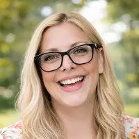 Eine lächelnde Frau mit langen blonden Haaren und schwarzer Brille steht im Freien, mit grünen Bäumen und Sonnenlicht im unscharfen Hintergrund - vielleicht eine fachkundige Mitarbeiterin der Immobilien Schenkung Notar Berlin.