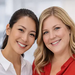 Zwei eng beieinander lächelnde Frauen, eine mit langen dunklen Haaren in einem Pferdeschwanz und weißer Bluse, die andere mit schulterlangen blonden Haaren und roter Bluse, stehen in einem hellen Innenraum und besprechen möglicherweise Angelegenheiten der GmbH Verkauf Notar.