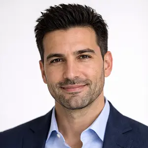 Ein Mann mit kurzen dunklen Haaren und leichten Bartstoppeln lächelt leicht. Er trägt ein hellblaues Hemd und einen marineblauen Blazer und steht vor einem schlichten weißen Hintergrund - ideal für ein professionelles Profil von Geschäftsführerwechsel Notar Berlin.