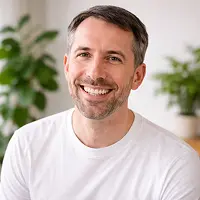 Ein lächelnder Mann mit kurzen braunen Haaren und einem Bart, der ein weißes T-Shirt trägt, sitzt in einem Innenraum mit Grünpflanzen und sanftem Licht im Hintergrund und wirkt entspannt, nachdem er die Nachricht über seine Grundschuldlöschung erhalten hat.