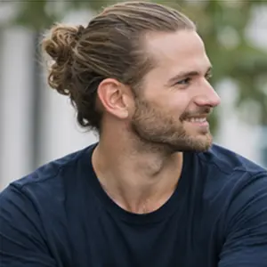 Ein Mann mit langen braunen Haaren, die zu einem Dutt gebunden sind, und einem Bart lächelt und blickt zur Seite, vielleicht inspiriert von den Plänen zu UG gründen. Er trägt ein dunkles Hemd, und der Hintergrund ist unscharf mit Grünpflanzen.