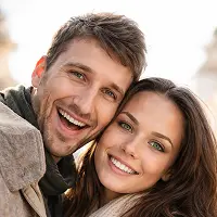 Ein lächelndes junges Paar im Freien, Wange an Wange. Der Mann hat kurzes braunes Haar und Bartstoppeln, die Frau hat langes dunkles Haar. Beide haben ein strahlendes Lächeln und wirken glücklich - vielleicht feiern sie mit ihrem Immobilien-Notar in Berlin.