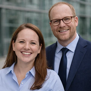 Eine lächelnde Frau mit braunen Haaren und einem hellblauen Hemd steht neben einem Mann mit Brille, Bart und blauem Anzug und Krawatte. Beide stehen im Freien vor einem Glasgebäude und treffen sich möglicherweise mit ihrem Notar, um eine Umfirmierung zu besprechen.