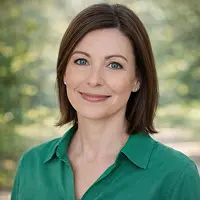 Eine Frau mit glattem braunem Haar und blauen Augen lächelt sanft in die Kamera. Sie trägt ein grünes Hemd mit Kragen und spricht vielleicht über Grundschuldlöschung, während sie im Freien vor einem unscharfen Hintergrund aus Grünzeug steht.