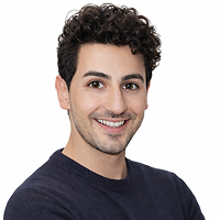 Ein junger Mann mit lockigem dunklem Haar und heller Haut lächelt in die Kamera. Er trägt ein dunkles langärmeliges Hemd und steht vor einem schlichten weißen Hintergrund.