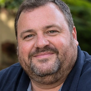 Ein Mann mit kurzen braunen Haaren und einem Bart lächelt in die Kamera. Bekleidet mit einem dunkelblauen Hemd steht er im Freien inmitten von verschwommenem Grün und denkt vielleicht über Fragen wie "Was kostet eine Gründung" in seinem Alltag nach.