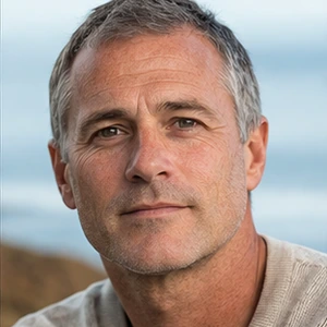 Ein Mann mittleren Alters mit kurzen grauen Haaren und leichten Bartstoppeln trägt einen beigen Pullover. Er blickt direkt in die Kamera mit einem neutralen Gesichtsausdruck, als würde er für ein Musterprotokoll posieren, mit einem unscharfen Außenhintergrund hinter ihm.