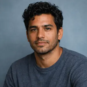 Ein Mann mit kurzen lockigen dunklen Haaren und Bartstoppeln, der ein graues T-Shirt mit Rundhalsausschnitt trägt, posiert vor einem schlichten blauen Hintergrund und blickt ruhig in die Kamera - wie jemand, der bereit ist, Stammkapital für eine GmbH oder UG einzuzahlen.