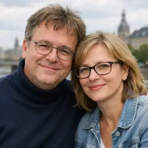 Ein lächelnder Mann und eine Frau mit Brille posieren eng beieinander im Freien, mit verschwommenen Gebäuden und einem Fluss im Hintergrund. Der Mann trägt einen dunklen Pullover, die Frau eine Jeansjacke - vielleicht besprechen sie Eigentümergrundschuld-Angelegenheiten.