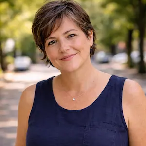 Eine Frau mit kurzen braunen Haaren, die ein dunkles ärmelloses Oberteil trägt, lächelt sanft im Freien auf einer von Bäumen gesäumten Straße und hält vielleicht inne, um notarielle Entwürfe zu prüfen; der Hintergrund ist weich und unscharf.