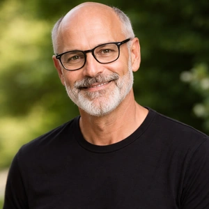 Ein lächelnder älterer Mann mit grauem Bart und Brille, der ein schwarzes T-Shirt trägt, steht im Freien mit grünem Laub im Hintergrund - vielleicht genießt er einen ruhigen Moment, nachdem er seinen Notar Erbrecht Berlin konsultiert hat.