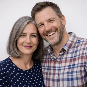 Eine lächelnde Frau mit grauen Haaren in einer marineblauen gepunkteten Bluse steht neben einem Mann in einem karierten Hemd; beide sehen glücklich aus und stehen eng beieinander - wie mehrere Gesellschafter, die bereit sind, UG zu gründen - vor einem schlichten Hintergrund.