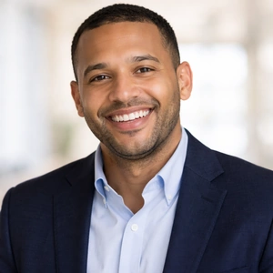 Ein Mann in einem marineblauen Blazer und einem hellblauen Hemd lächelt in die Kamera. Sein kurzes Haar und sein gestutzter Bart verleihen ihm ein gepflegtes Aussehen. Der leicht verschwommene Hintergrund deutet auf einen Innenraum hin, vielleicht perfekt für diejenigen, die an Online-Gründertreffen interessiert sind.