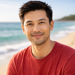 Ein junger Mann mit kurzen dunklen Haaren und gestutztem Bart lächelt in die Kamera, während er in einem roten T-Shirt an einem Sandstrand sitzt. Im Hintergrund sind das Meer, der blaue Himmel und sein Laptop zu sehen, auf dem Online Notar geöffnet ist.
