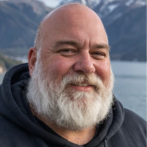 Ein bärtiger Mann mit Glatze lächelt im Freien und trägt einen dunklen Kapuzenpulli - vielleicht träumt er davon, wie er die Gesellschaft aufbauen kann. Im Hintergrund sind schneebedeckte Berge und ein Gewässer zu sehen.