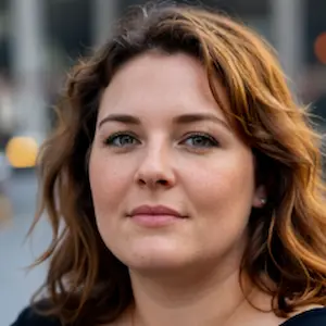 Eine Frau mit gewelltem, schulterlangem braunem Haar und heller Haut schaut im Freien direkt in die Kamera. Sie hat grüne Augen und einen neutralen Gesichtsausdruck, als ob sie darüber nachdenkt, wie sie satzung erstellen soll. Der unscharfe Hintergrund deutet auf eine städtische Umgebung hin.