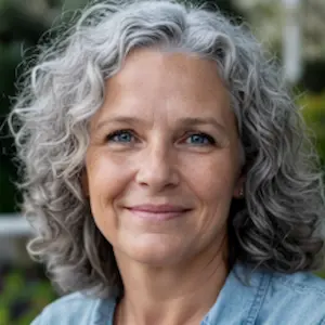 Eine Frau mittleren Alters mit lockigen grauen Haaren und blauen Augen, die Rechtsanwältin für Zivilrecht ist, lächelt sanft in die Kamera. Sie steht im Freien, das Grün im Hintergrund ist verschwommen, und trägt ein hellblaues Hemd.
