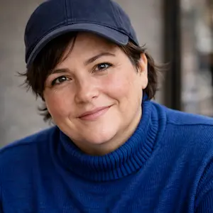 Eine Person, die eine dunkelblaue Baseballmütze und einen blauen Rollkragenpullover trägt, lächelt sanft in die Kamera und denkt vielleicht über die Rollen und Aufgaben der Gesellschafter nach. Der Hintergrund ist sanft verschwommen.