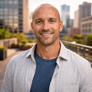 Ein lächelnder Mann mit rasiertem Kopf und gestutztem Bart trägt ein blaues T-Shirt und ein hellgraues Button-up-Hemd und steht im Freien auf dem Dach einer Stadt. Als Rechtsanwalt in Berlin genießt er die städtische Landschaft mit Gebäuden und Grünflächen im Hintergrund.