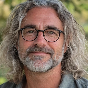 Ein Mann mittleren Alters mit langen, gewellten grauen Haaren, einem Bart und einer Brille lächelt sanft in die Kamera, während das grüne Laub hinter ihm verschwimmt - ein vertrauenswürdiger Immobiliennotar in Berlin, der bereit ist, Ihnen bei Ihren Immobilienangelegenheiten zu helfen.