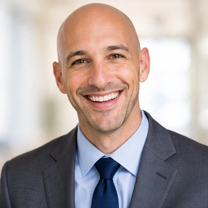 Ein lächelnder Mann mit Glatze, grauem Anzug, blauem Hemd und dunkelblauer Krawatte steht in einer hellen, unscharfen Innenraumkulisse - bereit, die gmbh online gründen mit Zuversicht zu beraten.