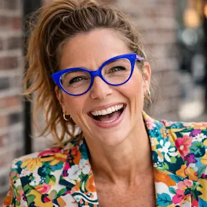 Eine Frau mit gewelltem blondem Haar in einem Pferdeschwanz, die eine hellblaue Brille und einen bunten geblümten Blazer trägt, lächelt breit vor einem unscharfen Backsteinmauer-Hintergrund und strahlt ein Selbstbewusstsein aus, das den Vertretungsregelungen des Geschäftsführers entspricht.