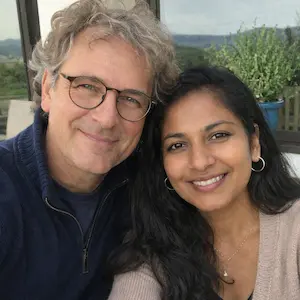 Ein lächelnder Mann mit Brille und grauem Haar sitzt neben einer lächelnden Frau mit langen dunklen Haaren. Sie sind im Freien, genießen das Grün und die Hügel im Hintergrund, vielleicht feiern sie gemeinsam den Erhalt ihrer Löschungsbewilligung.